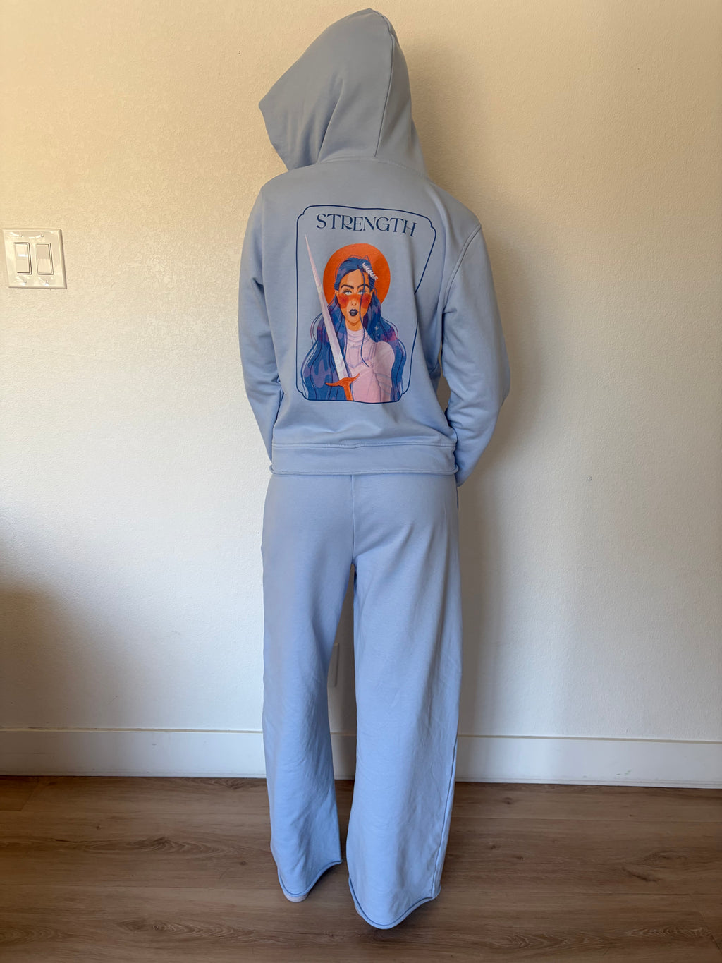 A person standing indoors wearing a light blue hooded sweatshirt and matching sweatpants, shown from the back. The hoodie has a large graphic print on the back featuring a stylized religious figure with the word 'STRENGTH' above it.