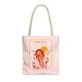 A light pink canvas tote bag with beige handles, featuring a printed graphic of a woman with red hair under a yellow sun and the text 'THE SUN'.