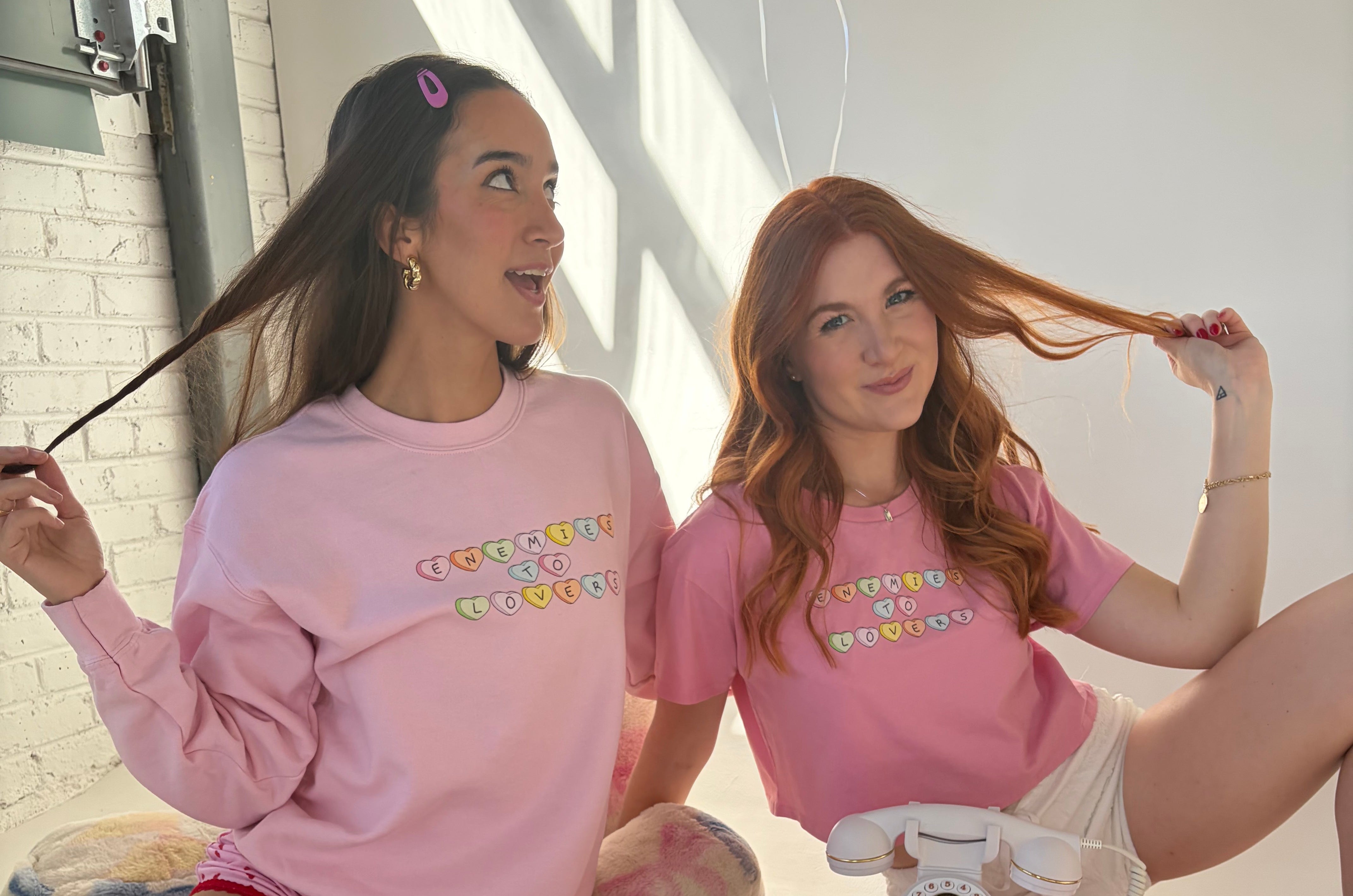 Two girls wearing enemies to lovers sweatshirts smiling with a popcorn bowl in front