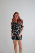 A woman with pink hair stands against a plain light gray wall wearing a short-sleeve collared romper with a black background and a colorful all-over print of small cartoon faces.