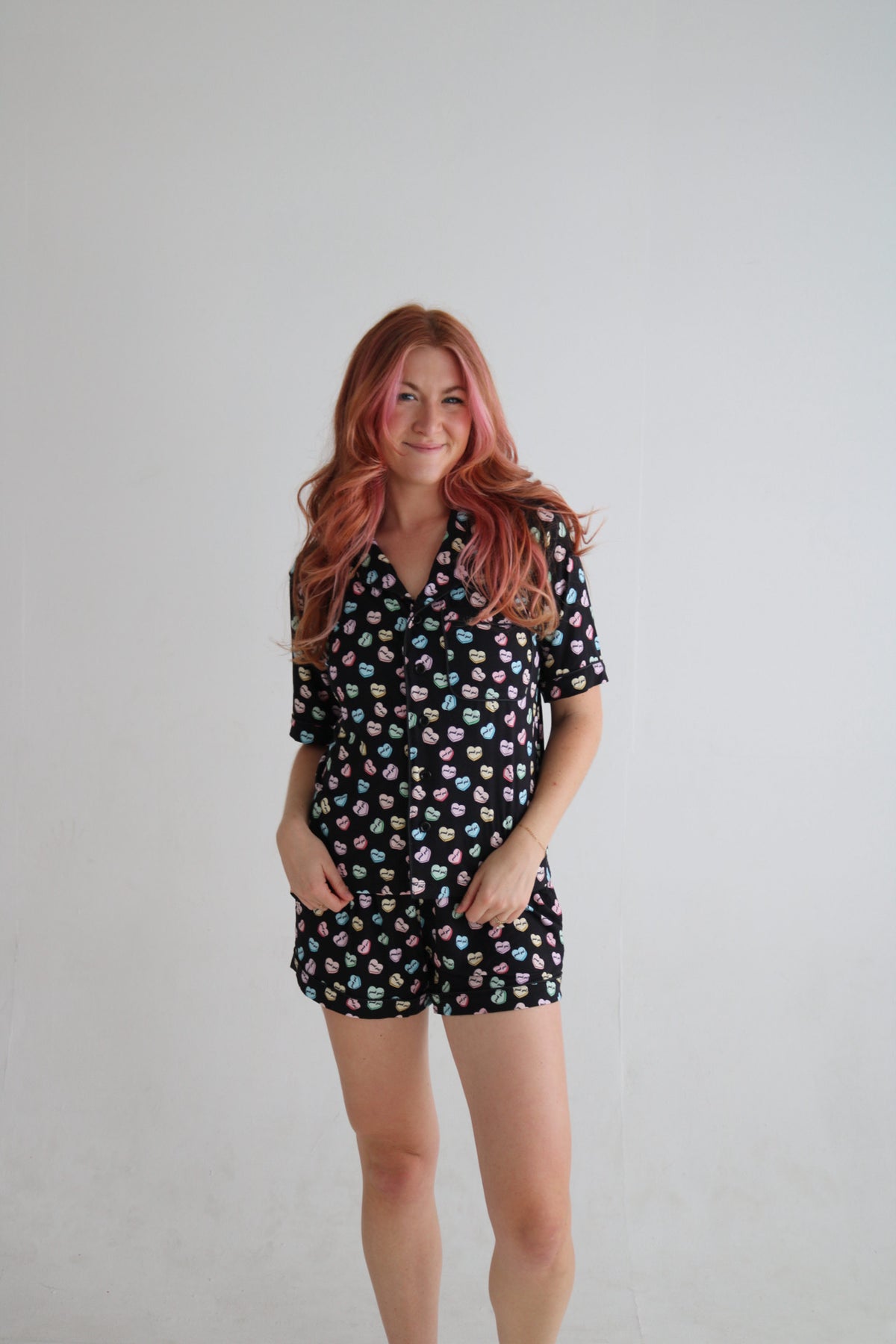 A woman with pink hair stands against a plain light gray wall wearing a short-sleeve collared romper with a black background and a colorful all-over print of small cartoon faces.