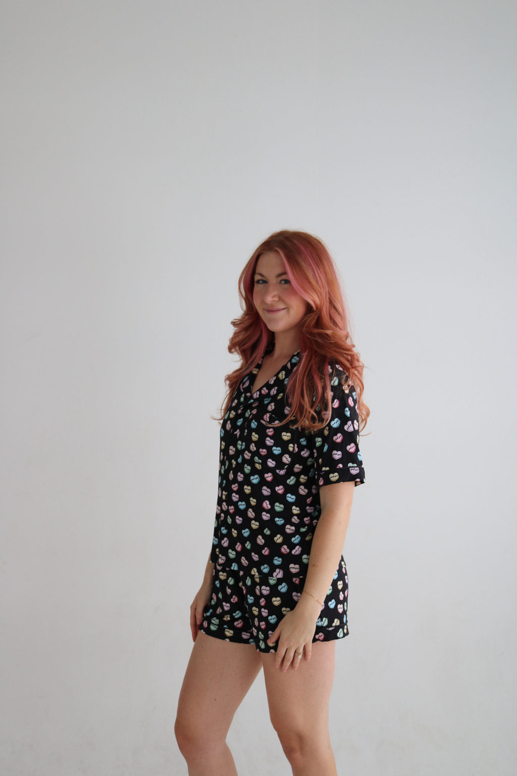 A woman with red hair wearing a short-sleeve black pajama romper with a colorful donut print, standing against a plain light gray background.