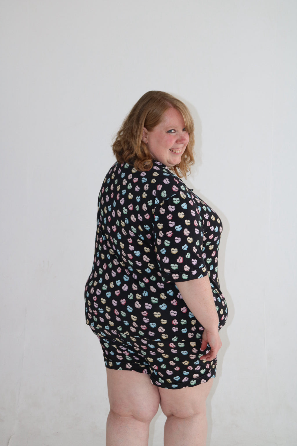 A woman modeling a black short-sleeve romper with a colorful pastel donut print, photographed against a plain white wall.