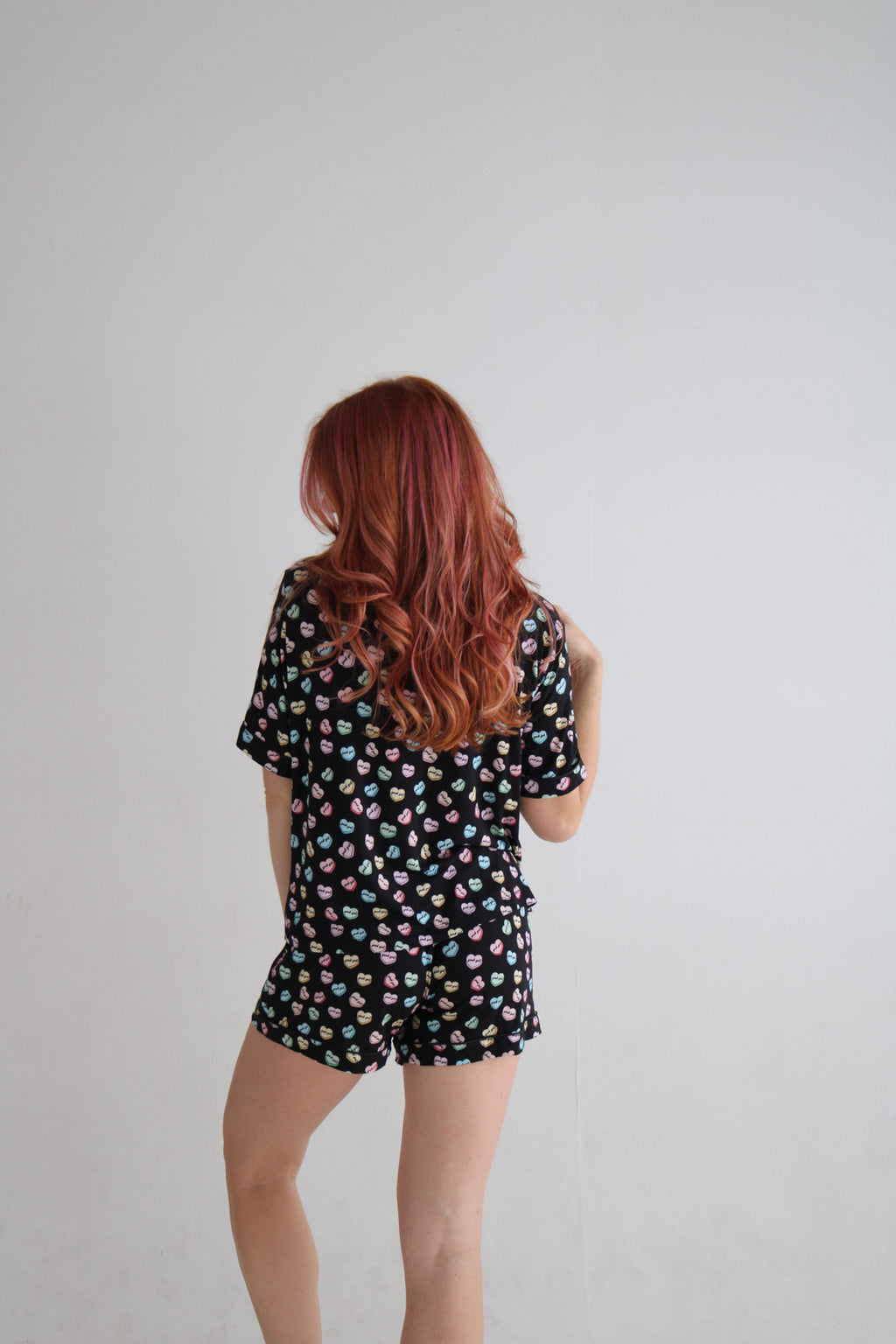 A woman with long red hair is shown from behind wearing a black short-sleeve romper with a colorful polka dot pattern, set against a plain light gray background.