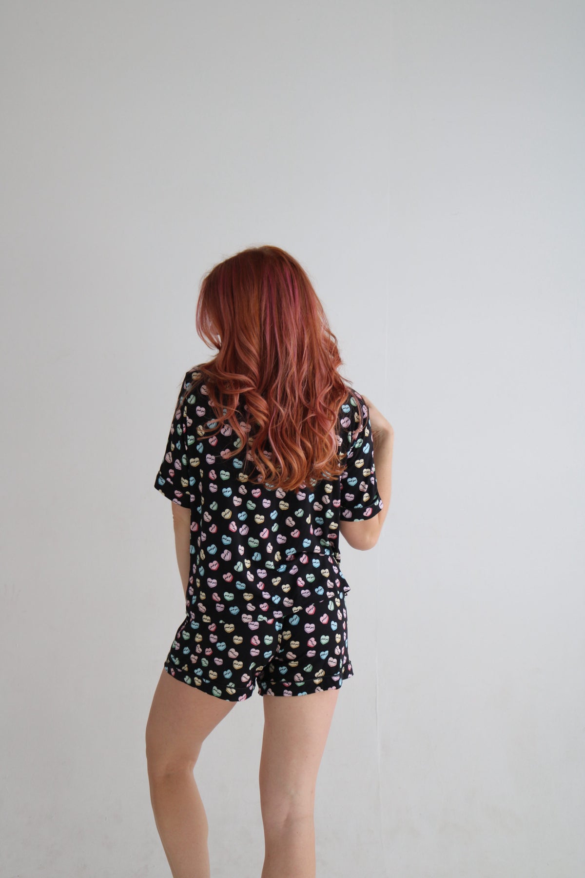 A woman with long red hair is shown from behind wearing a black short-sleeve romper with a colorful polka dot pattern, set against a plain light gray background.