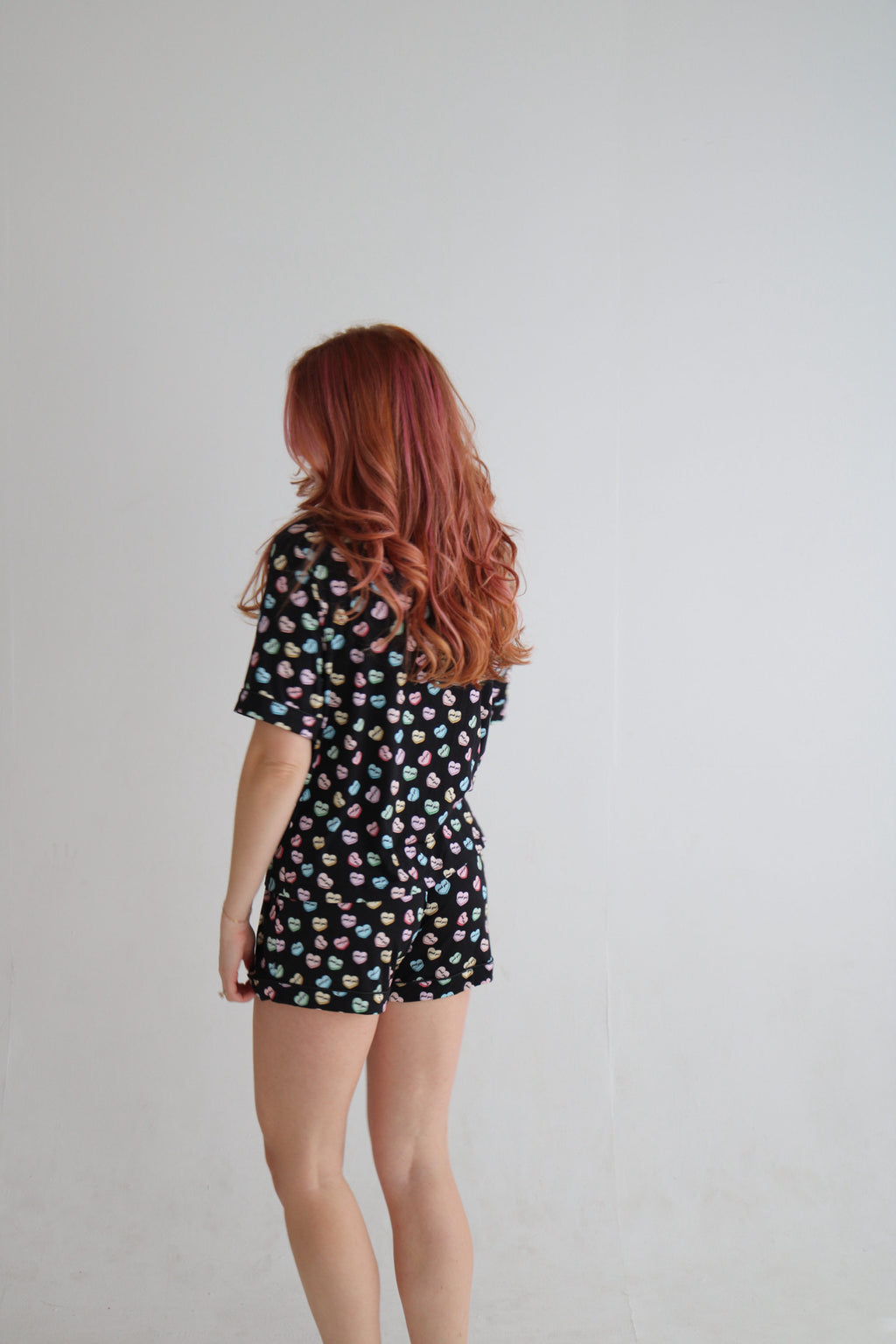 A woman with long wavy red hair is shown from behind wearing a short-sleeve romper with a black base and a colorful polka dot pattern, standing against a plain light gray wall.