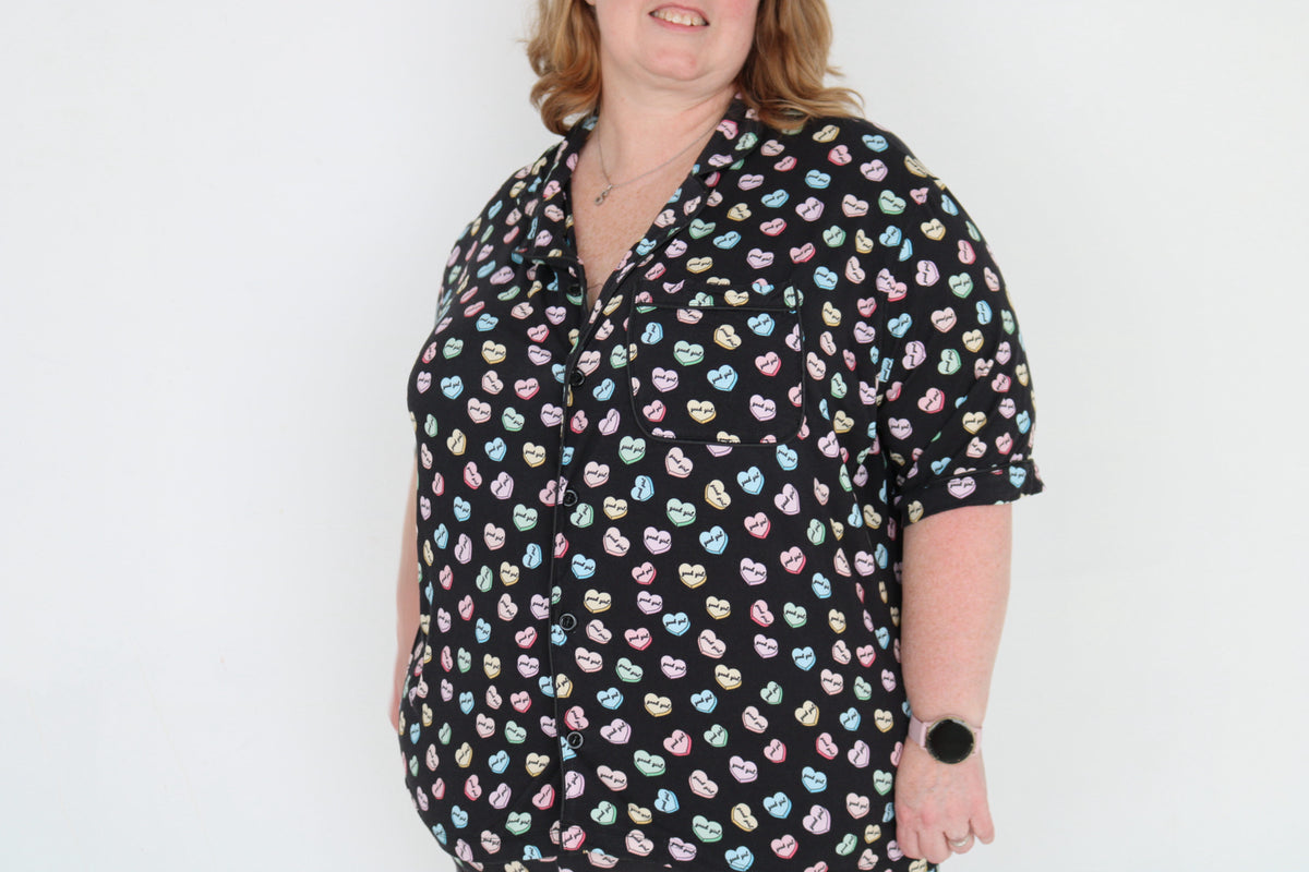 A woman wearing a black short-sleeve button-up shirt with a colorful pattern of small hearts, stars, and flowers, shown against a plain white background.