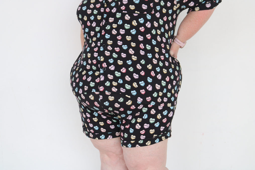 A person wearing a black romper with a colorful repeating pattern of small hearts, shown from the back against a plain white background.