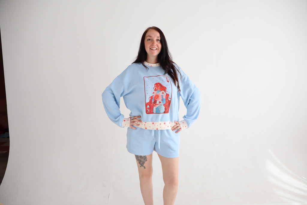 A woman modeling a light blue pajama set consisting of a long-sleeve sweatshirt with a red graphic print on the front and matching shorts, styled with a patterned waist tie, against a white studio backdrop.