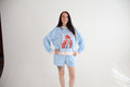 A woman modeling a light blue pajama set consisting of a long-sleeve sweatshirt with a red graphic print on the front and matching shorts, styled with a patterned waist tie, against a white studio backdrop.