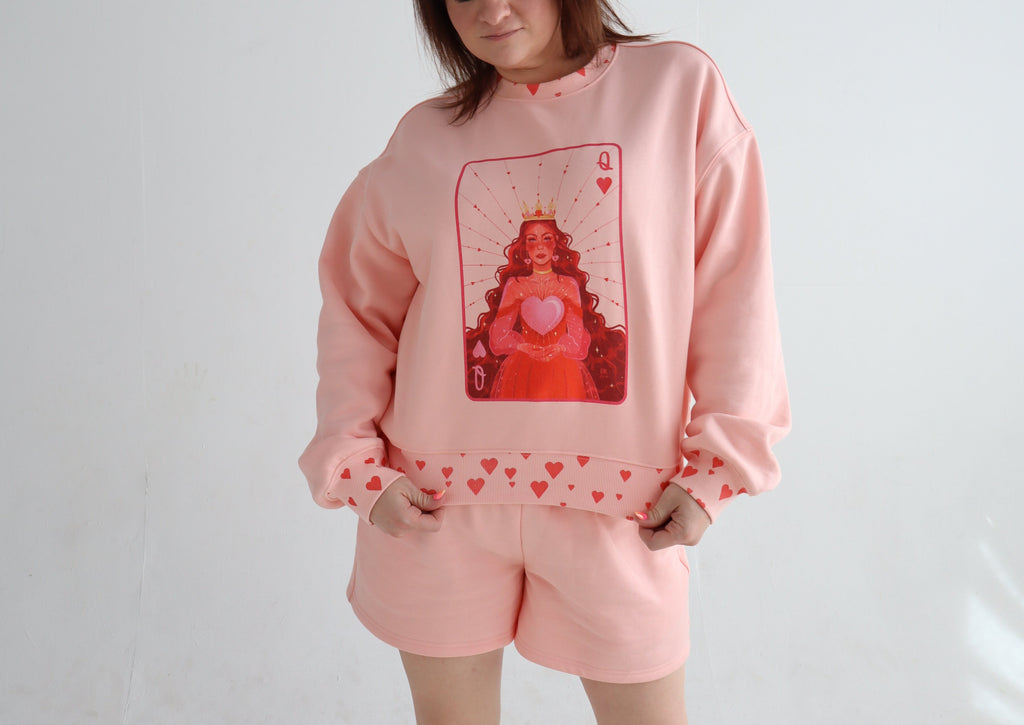 A woman modeling a light pink lounge set consisting of a sweatshirt and matching shorts, featuring a red playing card graphic on the sweatshirt and small red hearts along the hem.