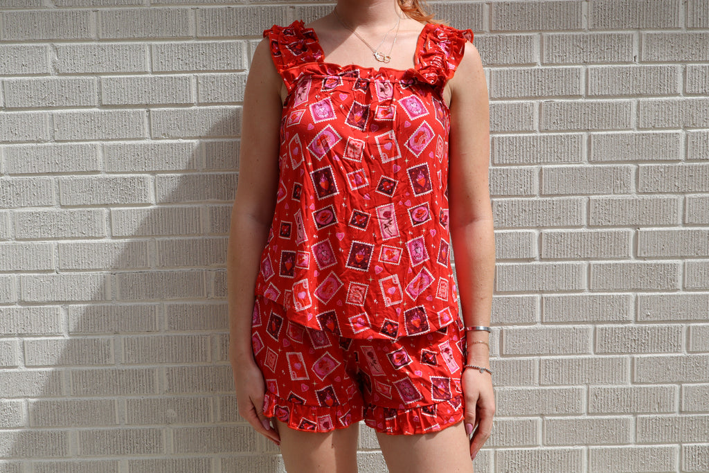 A woman wearing a red sleeveless romper with a pink and black geometric pattern, standing against a light gray brick wall.