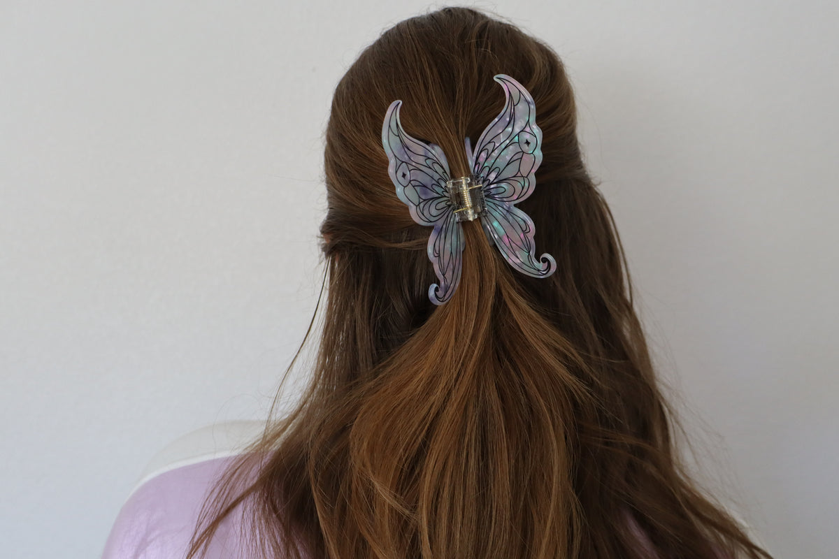 A butterfly-shaped hair claw clip with iridescent pastel wings, shown attached to a model's brown hair from the back against a plain light gray wall.