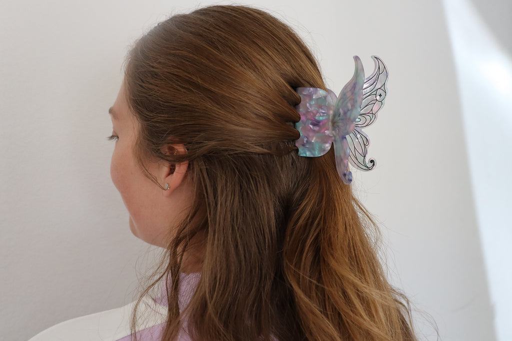 A woman with long brown hair shown from the side, wearing a translucent iridescent butterfly hair clip in a half-up ponytail.
