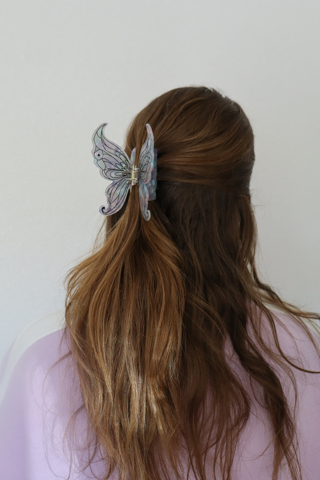 A woman viewed from behind with long brown hair, wearing a pastel butterfly hair clip on the side of her hair. She is wearing a light lavender top against a plain off-white background.