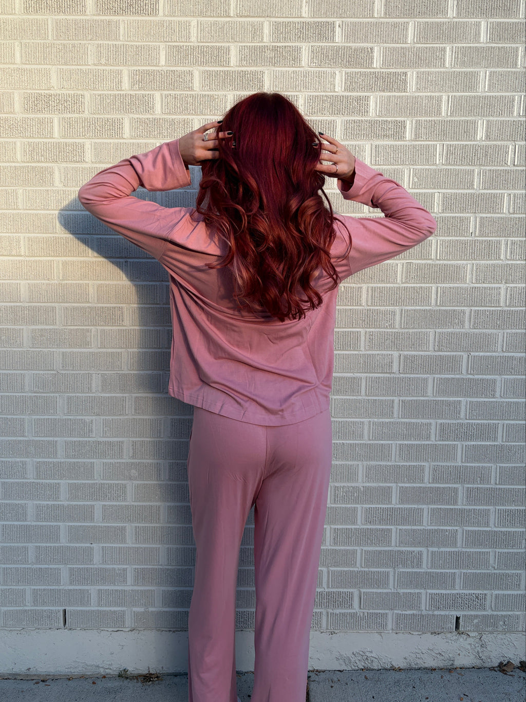 A person with long, wavy red hair stands with their back to the camera, wearing a matching dusty pink long-sleeve top and sweatpants set, posing against a light gray brick wall.