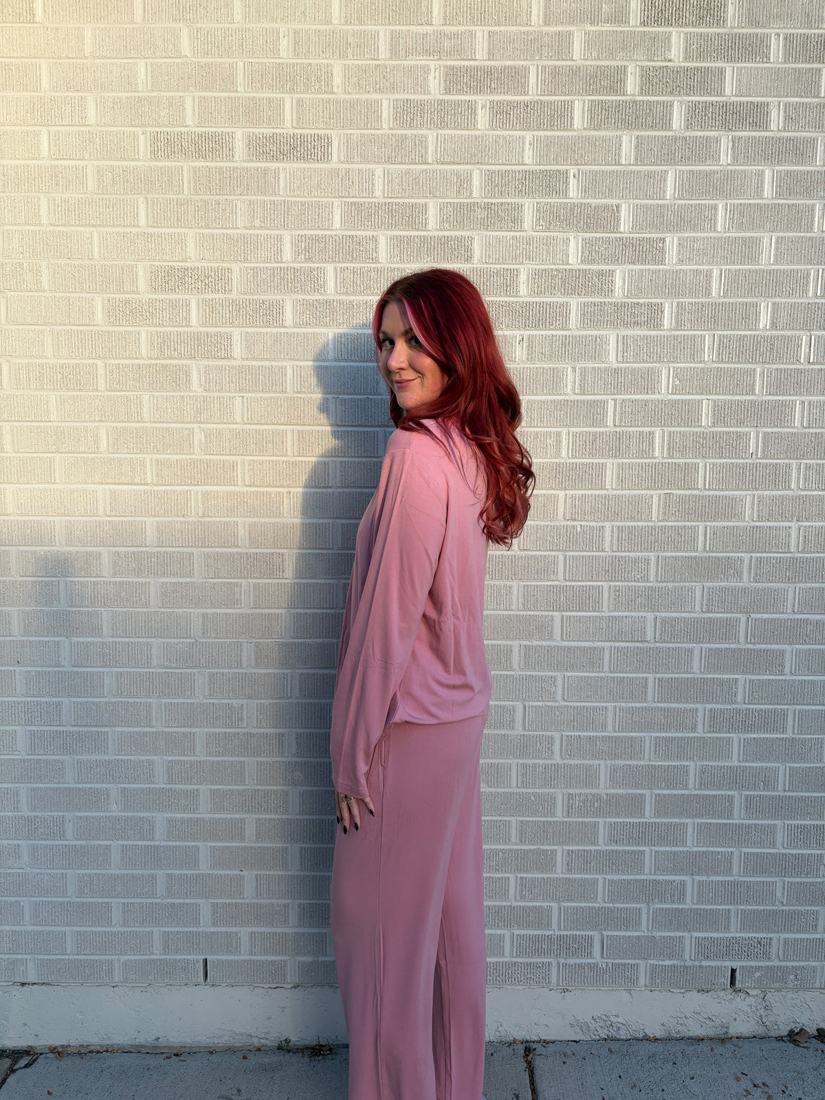 A woman with long red hair stands in profile against a light gray brick wall, wearing a matching dusty pink long-sleeve top and wide-leg skirt set.