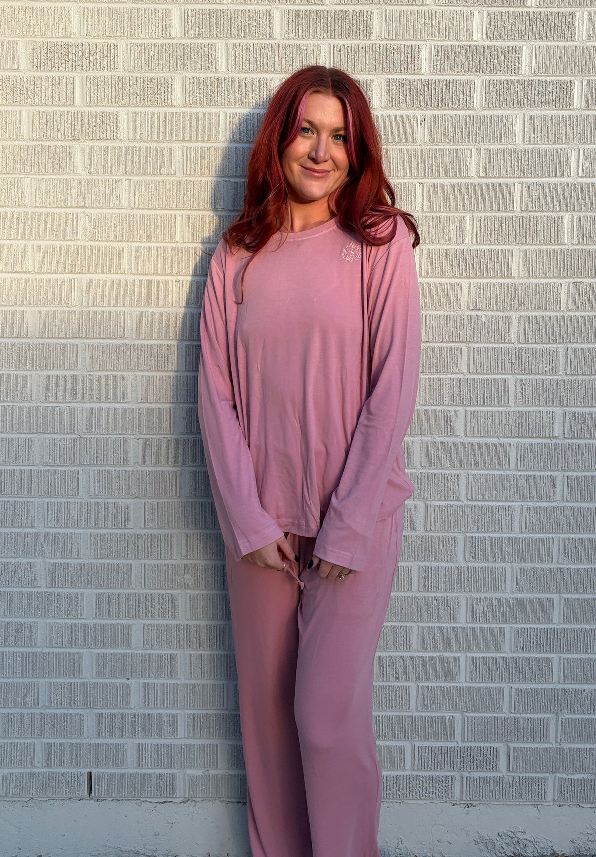 A woman with red hair stands against a light gray brick wall wearing a matching dusty pink lounge set consisting of a long-sleeve top and wide-leg pants.