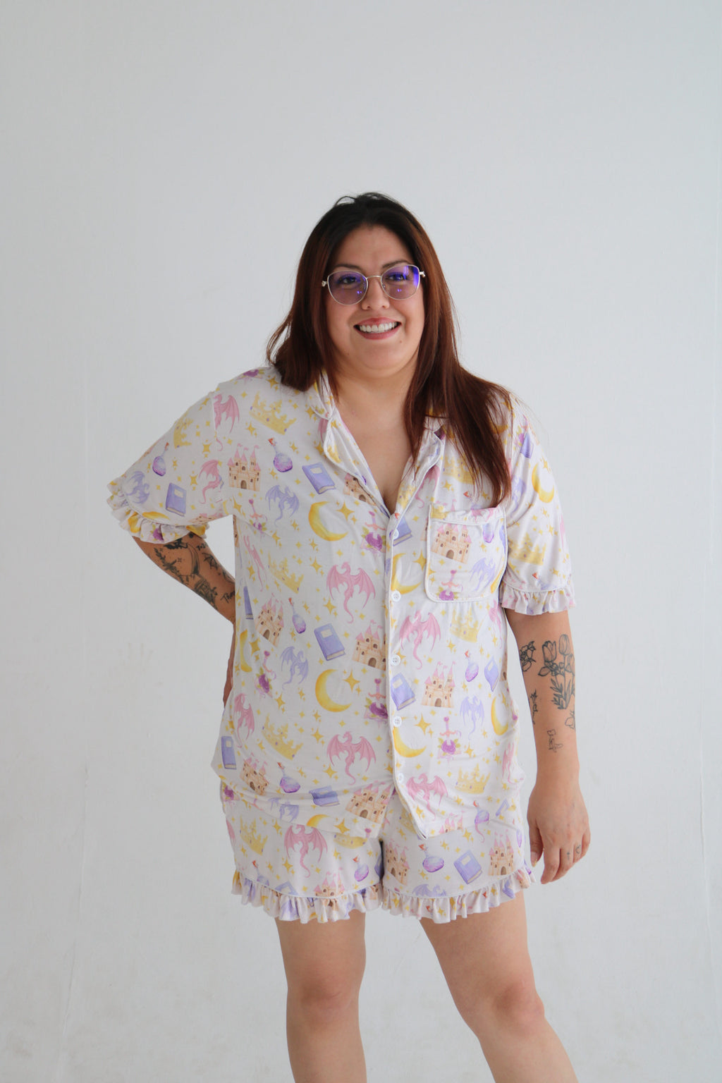 A woman modeling a matching short-sleeve button-up pajama shirt and fringed shorts set with a pastel celestial print of moons, stars, and clouds on a white background.