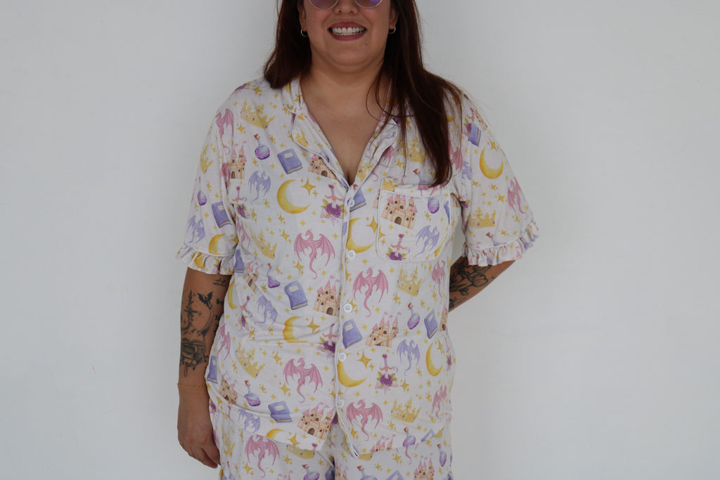 A woman wearing a matching short-sleeve pajama set with a whimsical pastel print of moons, stars, dragons, and flowers on a white background, standing against a plain light gray wall.