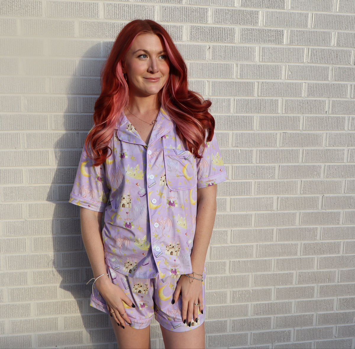 A woman with long red hair stands against a light gray brick wall wearing a light purple short-sleeve pajama set with a cartoon animal print.