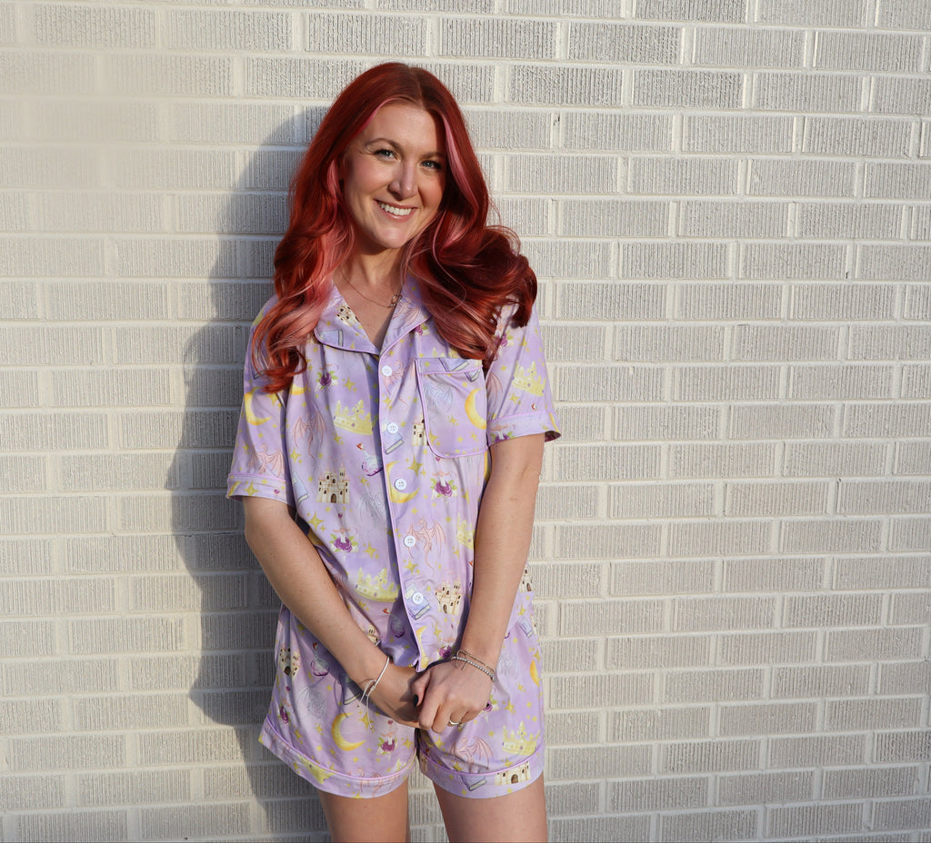 A woman with long red hair stands against a light gray brick wall wearing a lavender pajama set with a yellow and white crescent moon and floral pattern.