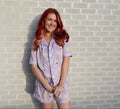A woman with long red hair stands against a light gray brick wall wearing a lavender pajama set with a yellow and white crescent moon and floral pattern.