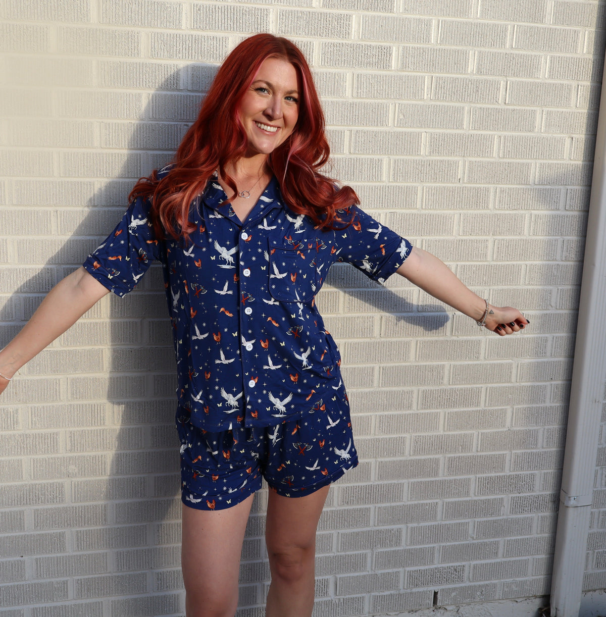 A woman with red hair wearing a navy blue short-sleeve button-up shirt and matching shorts set with a white and orange bird pattern, posing against a light gray brick wall.