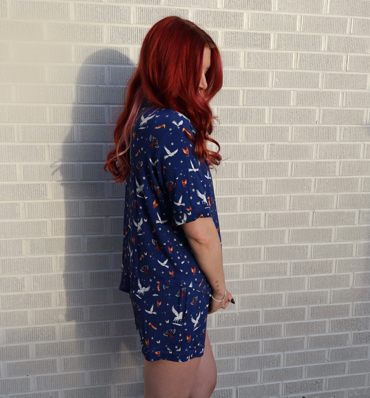 A woman with long red hair stands in profile against a white brick wall, wearing a navy blue short-sleeve pajama-style top and matching shorts set with a white bird and orange dot pattern.