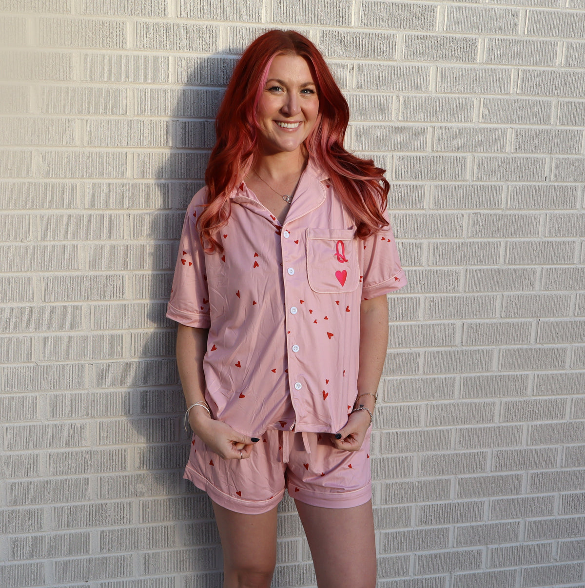 A woman with long red hair stands against a white brick wall wearing a light pink pajama set with small red heart prints.