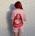 A person with red hair stands with their back to the camera, wearing a light pink pajama set with a large graphic of a woman in a crown on the back. The setting is outdoors against a white brick wall.