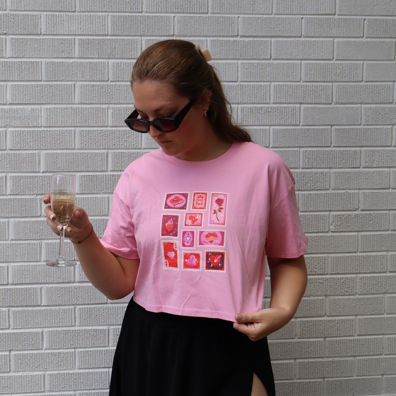 A woman wearing a pink short-sleeve crop top with a red graphic print and a black skirt, holding a glass of water in front of a gray brick wall.