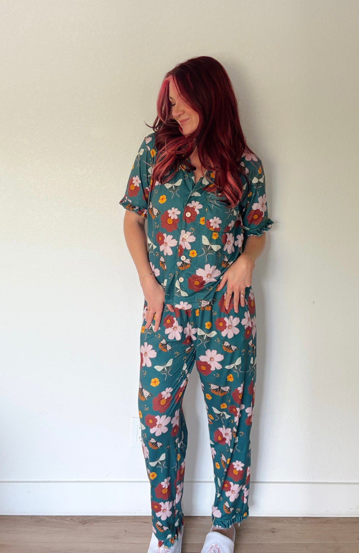 A woman with red hair wearing a matching short-sleeve button-up shirt and drawstring pants set in a teal green floral print, standing against a plain off-white wall.