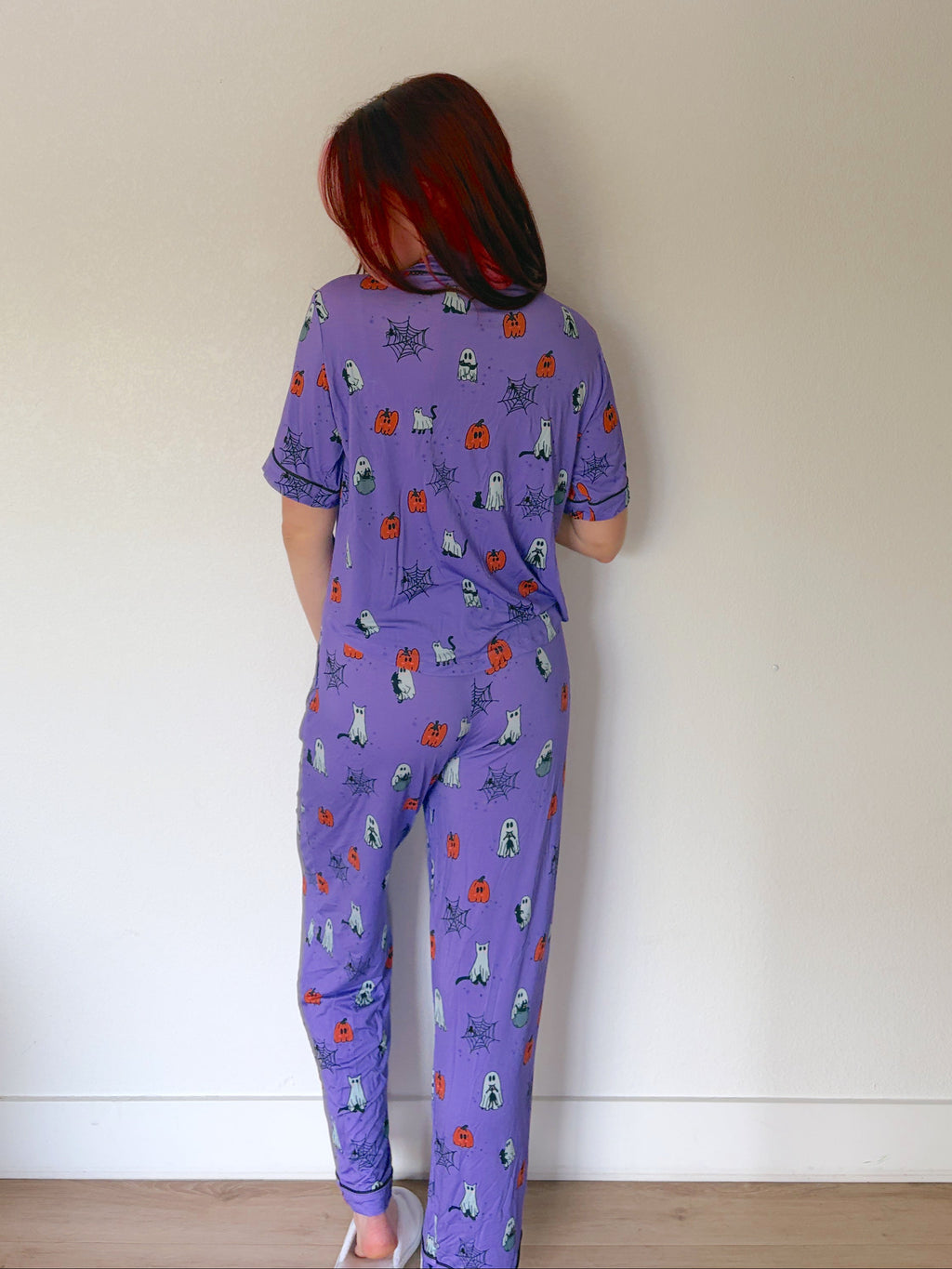 A person standing with their back to the camera wearing a purple short-sleeve pajama set with a small multicolor pattern, set against a plain off-white wall.