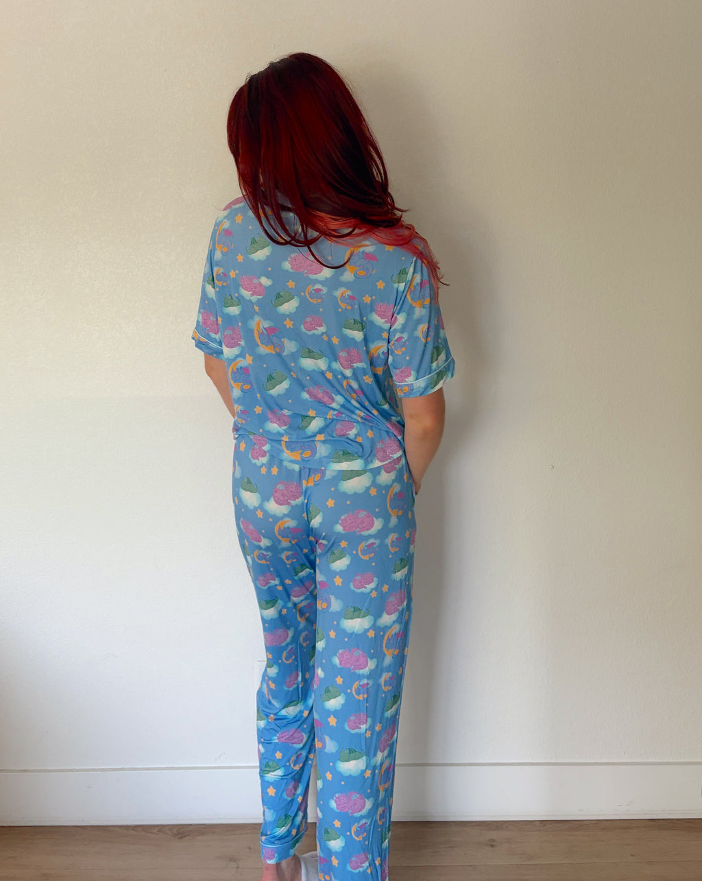 A person with red hair stands with their back to the camera wearing a matching blue short-sleeve pajama top and pants set with a pink and white mermaid pattern, set against a plain off-white wall.