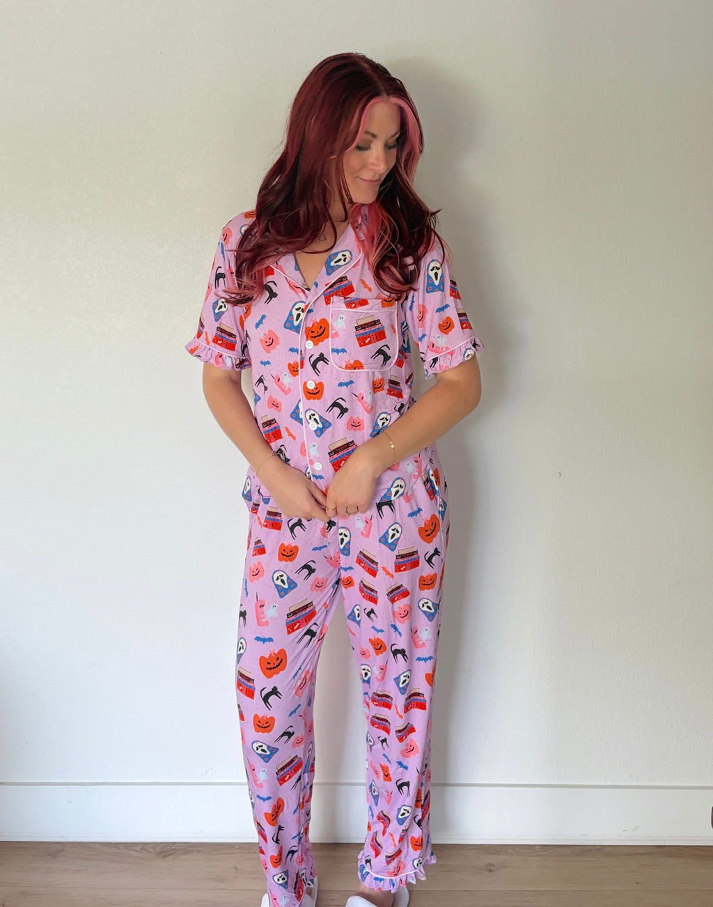 A woman with red hair wearing a pink short-sleeve pajama set with a colorful cartoon character print, standing against a plain off-white wall.