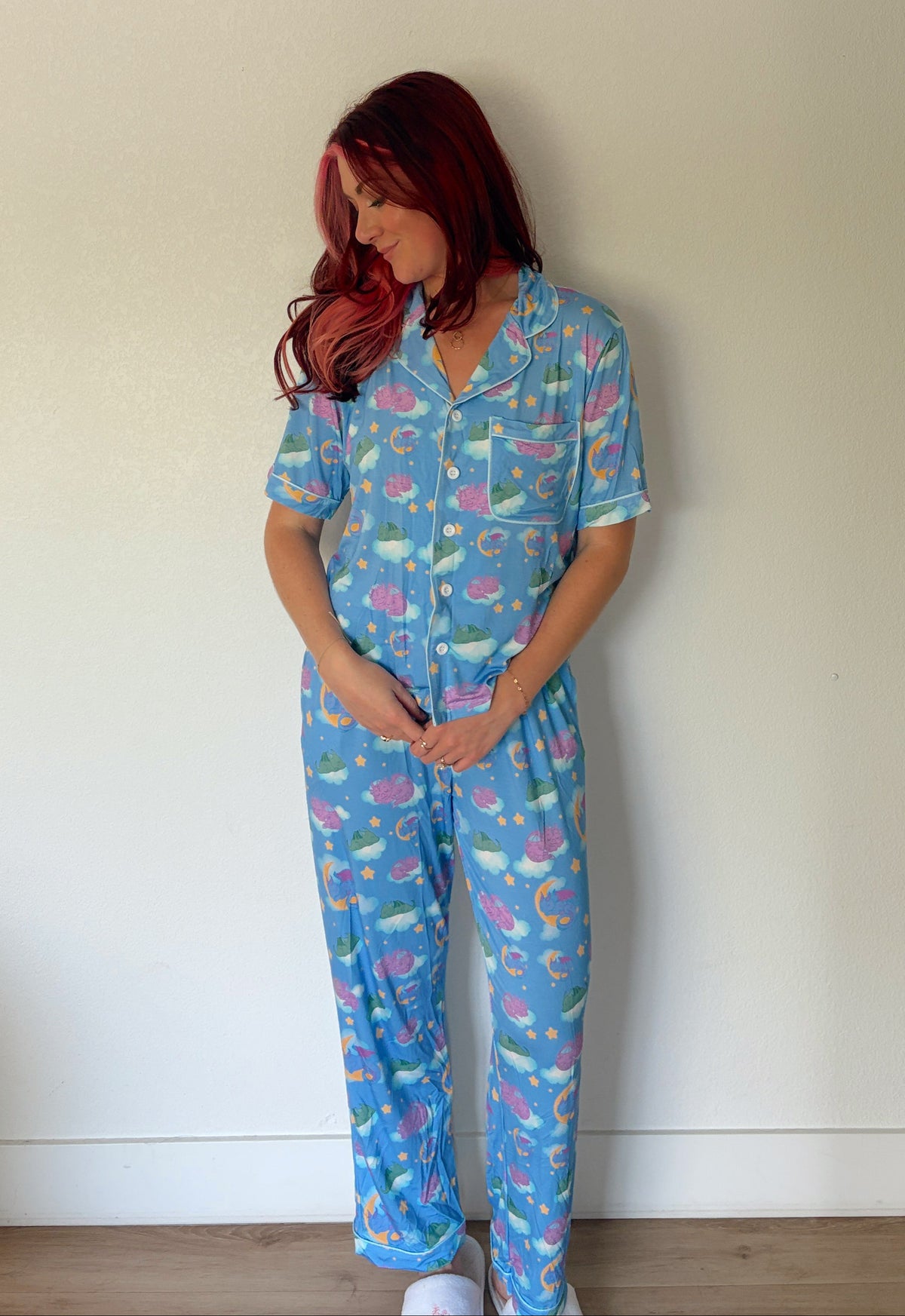A woman with red hair stands against a plain off-white wall wearing a light blue pajama set with a colorful tropical print of palm trees, flamingos, and sea life.