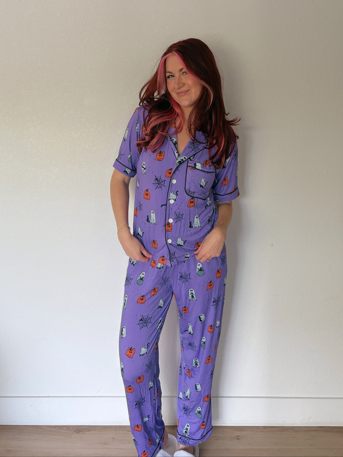 A woman wearing a purple short-sleeve pajama set with a cartoon print, standing against a plain white wall.