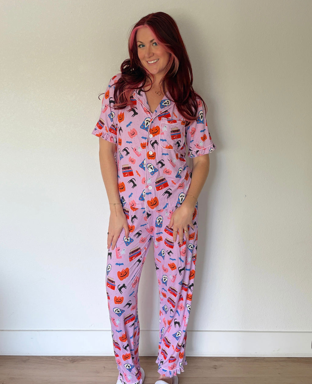 A woman with red hair wearing a pink short-sleeve pajama set with a colorful Halloween-themed print of pumpkins, ghosts, and bats, standing against a plain off-white wall.