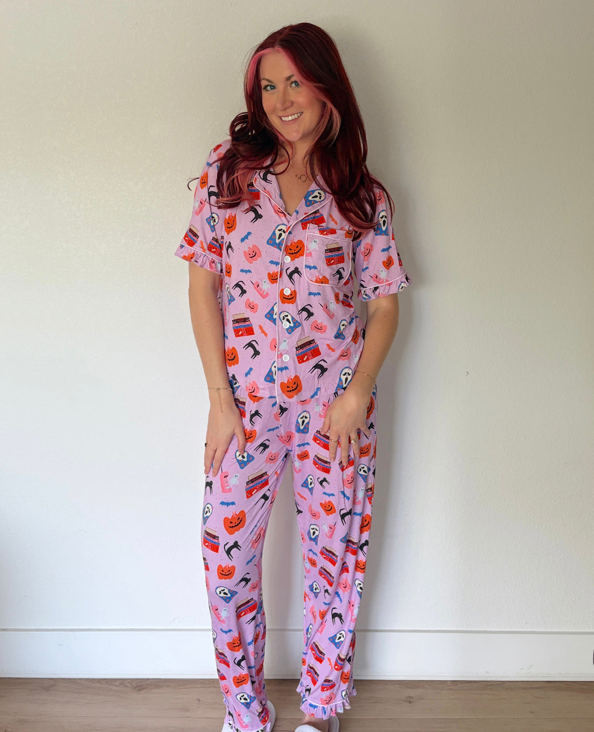 A woman with red hair wearing a pink short-sleeve pajama set with a colorful Halloween-themed print of pumpkins, ghosts, and bats, standing against a plain off-white wall.