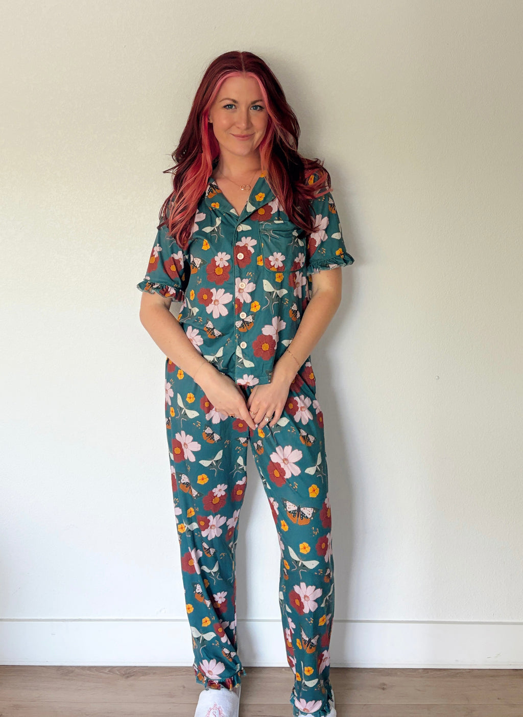 A woman with pink-tinted hair stands against a plain off-white wall wearing a matching short-sleeve button-up shirt and jogger pants set in a teal green floral print with white, pink, and orange flowers.
