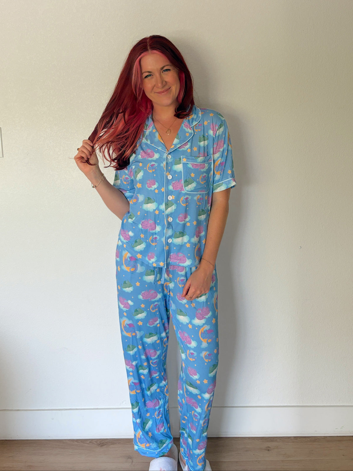 A woman with pink hair stands indoors wearing a light blue pajama set with a colorful mermaid pattern. The set includes a short-sleeve button-front top and matching long pants.