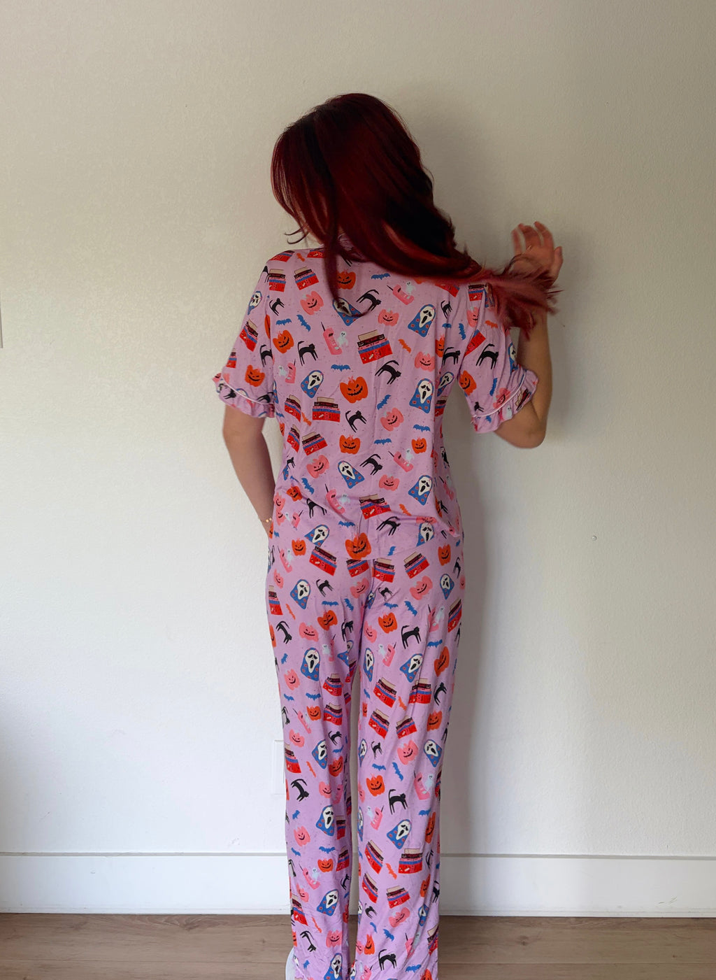A person with long red hair stands with their back to the camera wearing a pink short-sleeve scrub jumpsuit with a colorful medical-themed print, set against a plain white wall.