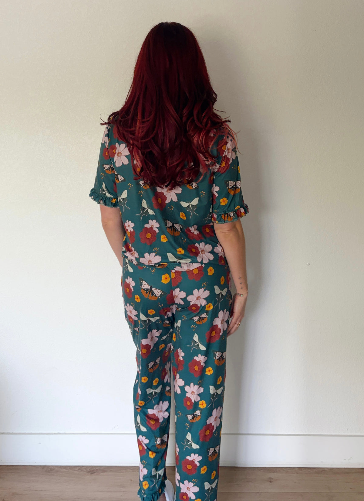 A woman with long, wavy dark red hair is shown from behind wearing a matching two-piece scrub set with a teal green background and a colorful floral print. The set includes a short-sleeve top and matching pants, photographed against a plain off-white wall.