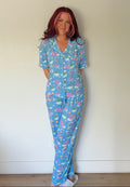 A woman with pink hair stands against a plain off-white wall wearing a light blue pajama set with a colorful tropical print of palm trees, suns, and fish.