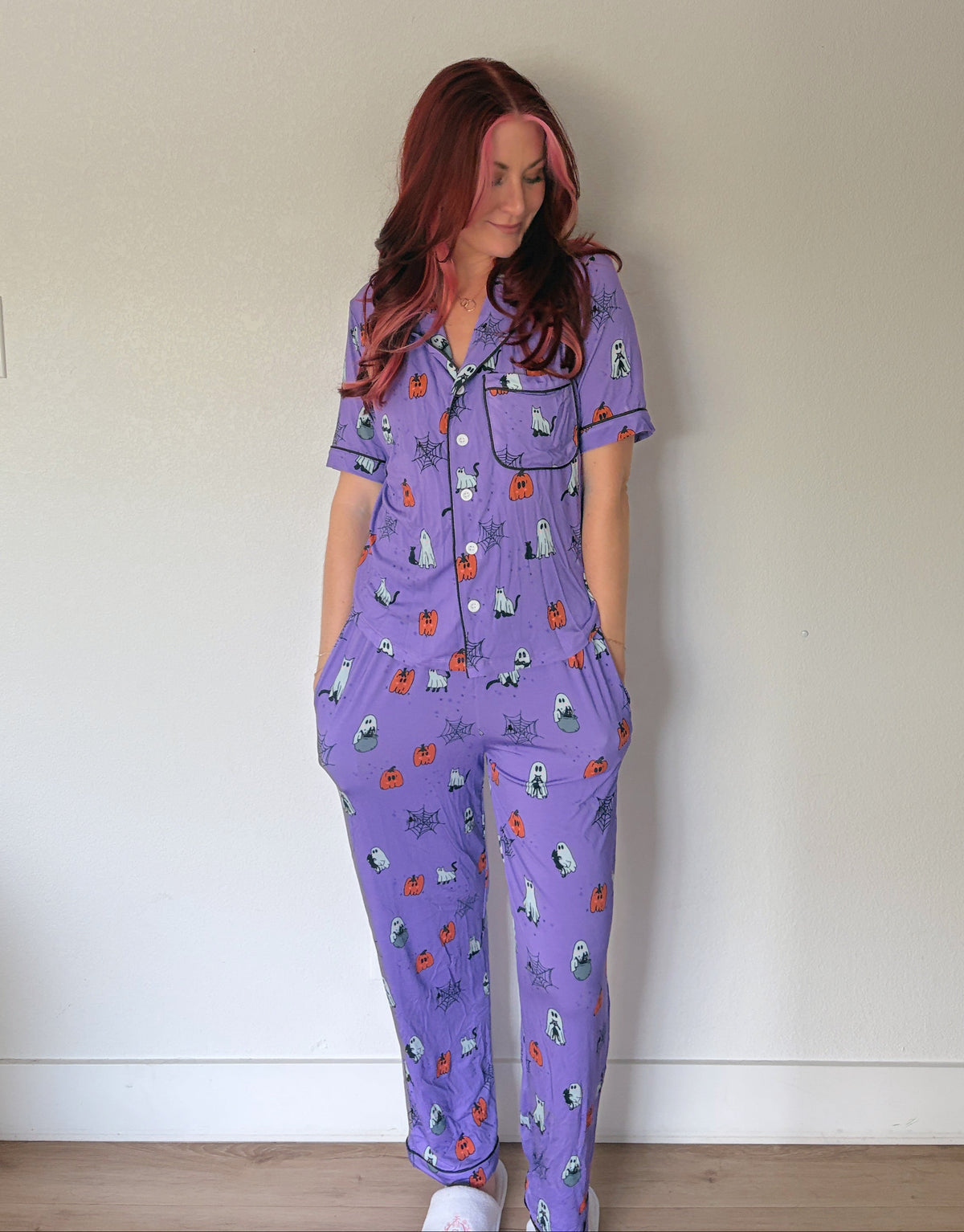 A woman with red hair stands against a plain off-white wall wearing a purple short-sleeve pajama set with a small cartoon print.