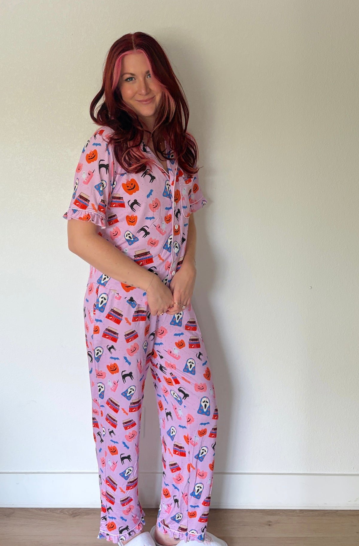 A woman with red hair wearing a pink short-sleeve pajama set with a colorful cartoon pattern, standing against a plain off-white wall.