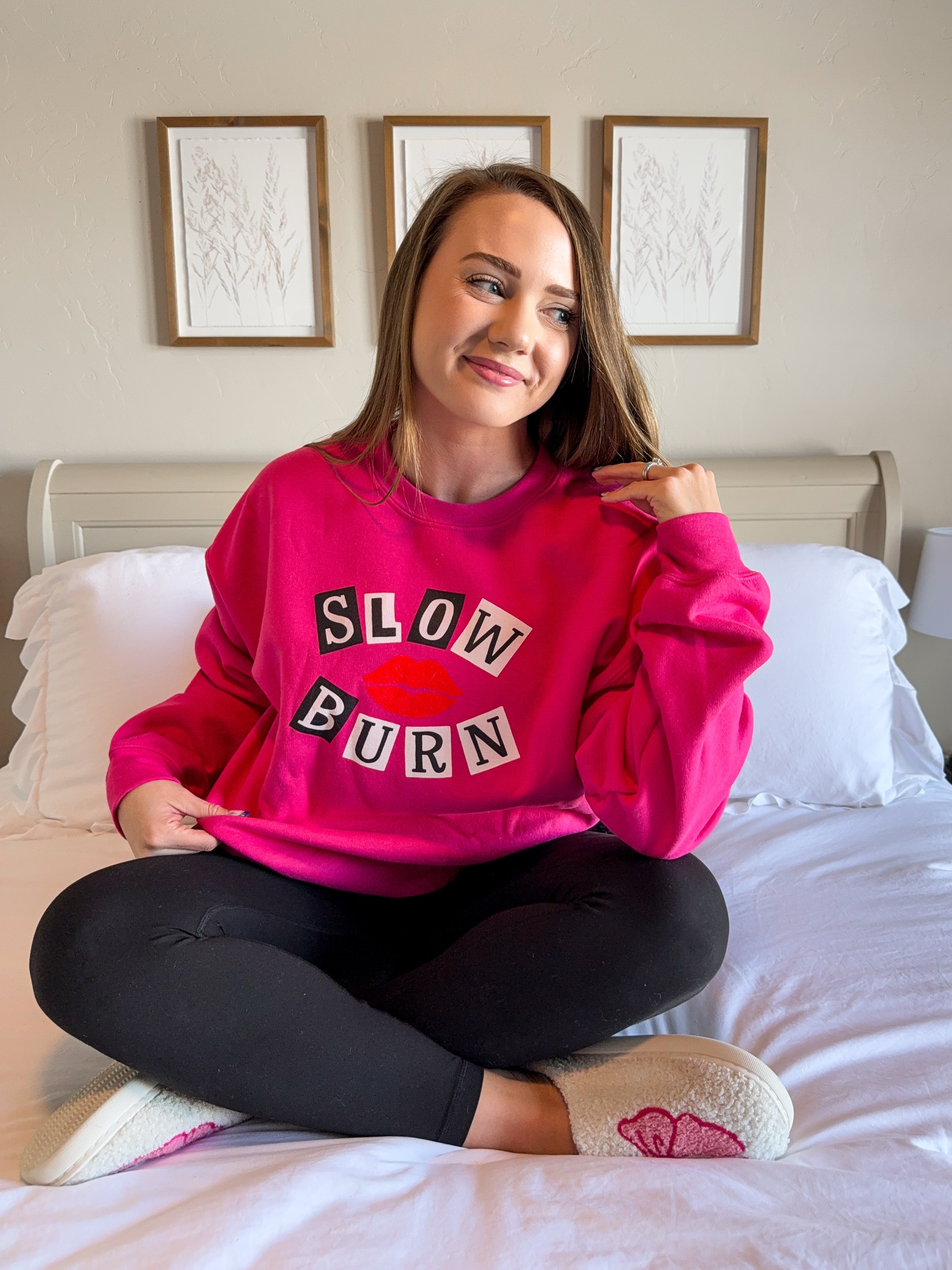 Woman wearing a pink sweatshirt with 'SLOW BURN' text, sitting on a bed.