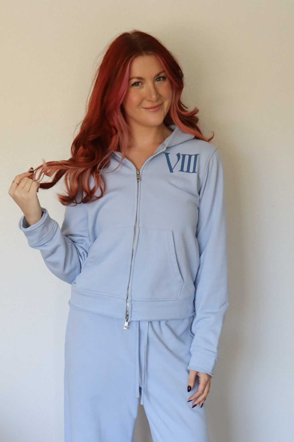 A woman with red hair wearing a light blue zip-up hoodie and matching wide-leg sweatpants with blue Roman numerals, standing against a plain off-white wall.